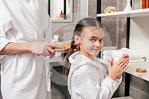 mother in white bathrobe combing daughter hair with wooden hairbrush near shelves