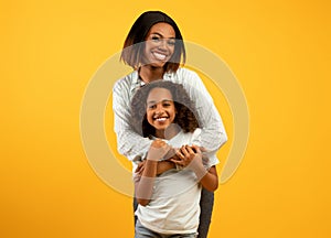 Mother& x27;s love. Happy loving african american mom cuddling her daughter from the back, standing over yellow background