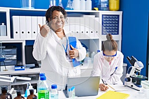 Mother and young daughter working at scientist laboratory pointing thumb up to the side smiling happy with open mouth