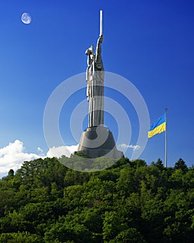 Motherland monument and waving ukrainian flag on green hill against blue sky in Kyiv, Ukraine