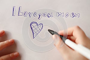Mothers day holiday.moms day. I love you mom inscription on a white sheet and a heart and a childs hand with a pen