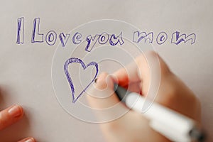 Mothers day.moms day.I love you mom inscription on a white sheet and a heart and a childs hand with a pen.