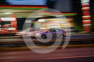 Motion shot of a gray car driving fast down a road near an illuminated gas station