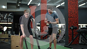 Motivated beginner sportsman hard time doing CrossFit workout with battle ropes by personal trainer standing with timer