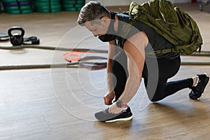 Motivated instructor kneeling and ties his shoestrings with backpack and training mask