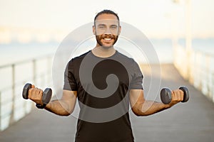 Motivated man strengthens arms lifting weights on sea pier outdoors