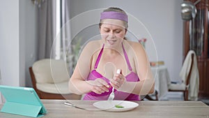 Motivated obese woman cutting cucumber slices with knife and eating healthful veggie food with fork. Portrait of young