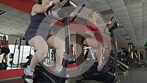 Motivated overweight and athletic women riding an exercise bike in the gym