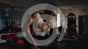 Motivated perspiring sportsman lifting dumbbell sitting in gym indoors. Medium shot portrait of sweating African