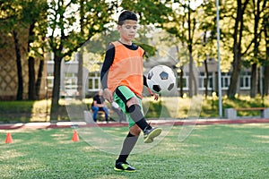 Motivated teen football player stuffs soccer ball on feet with boots. Practicing sport exercises at artificial stadium.