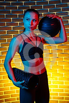 Motivated young brunette woman posing with medicine ball and foam fitness roller in gym in neon lights.