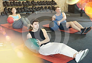 Motivated young man practicing exercises with medicine ball near other people in gym