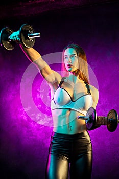 Motivated young woman fitness model exercise shoulders with professional dumbbells in neon lights in the studio.