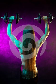 Motivated young woman fitness model exercise shoulders with professional dumbbells in neon lights in the studio. Back