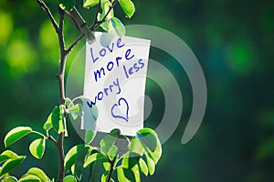 Motivating phrase love more worry less. On a green background on a branch is a white paper with a motivating phrase. photo