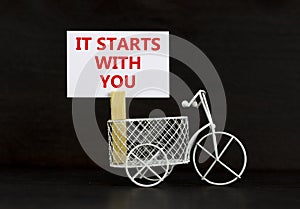 Motivational, start symbol. Wooden clothespin, white sheet of paper. Words `it starts with you`. Miniature bicycle model.