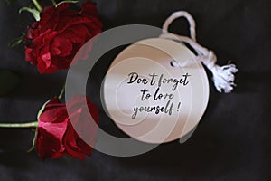 Motivational text message on white tag label paper - Do not forget to love yourself. With red roses on dark black background.