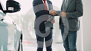 Motor showroom employee is giving car key to young male buyer and shaking hands, dealer is holding documents and
