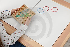 Motor skills development. Girl playing with geoboard and rubber bands at white table, closeup. Space for text