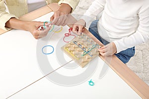 Motor skills development. Mother helping her daughter to play with geoboard and rubber bands at white table, closeup