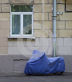 Motorcycle covered with a cover in bad weather stands under the window of an apartment building