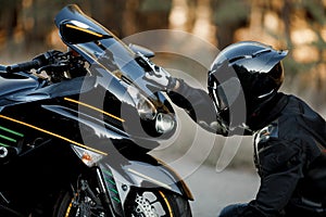 Motorcycle driver in leather suit and helmet sits in front of the motorcycle alone and put his hands on the headlights