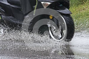 Motorcycle moped rides through a puddle on a wet road in the rain. Spray is flying from the wheels. Close-up