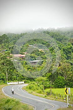 A motorcycle is running in the middle of a rural village road surrounded by mountains and fog in a refreshing morning.