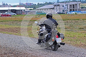 A motorcyclist in leather outfit rides a chopper on a dirt road along a dirt road against a backdrop of urban infrastructure. Hobb