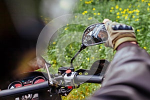 Motorcyclist looking in mirror of motobike outdoors. Man in leather jacket and helmet ride in summer forest