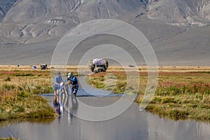 A motorcyclist pushes a motorcycle along a road flooded with water. In the background is a truck.Altai, Mongolia