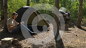 Motorcyclist sitting near tent in woods and finishing book at vacation near camp