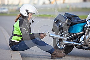 Motorcyclist woman typing a message in her smartphone while sitting on roadside near motorcycle