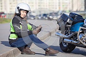 Motorcyclist woman in white crash helmet looking at camera, holding smartphone while sitting on roadside near chopper motorcycle