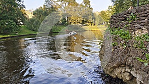 A motorized plastic boat floats on the river and creates a wave. One bank is reinforced with stone and the other with logs, on whi