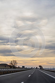 A motorway straight that gets lost against a cloudy dawn sky in vertical format, a car overtakes a truck further down the road