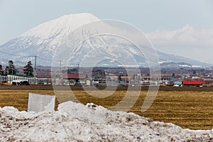 Mount Yotei inactive stratovolcano with village on the foot hill and yellow grass on the ground with pile of snow on foreground