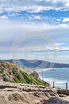 Mountain along shore of ocean with parachuters and pier against cloudy blue sky