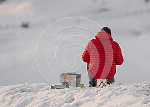 Mountain climber in red coat on white snow covered mountain hill taking break to keep warm eat and drink. Looking at hillside