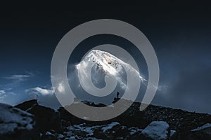 Mountain explorer on rock summit standing at night in front of snow cloudy high altitude mountain. Brave tourist on the top