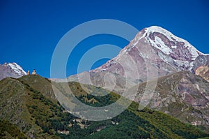 Mountain Kazbek with white snow covered peak and Gergeti Trinity church in the foreground with blue sky above