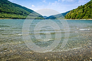 Mountain lake shore with waves in foreground, cloudy sky autumn day and distant green hills at horizon in the background