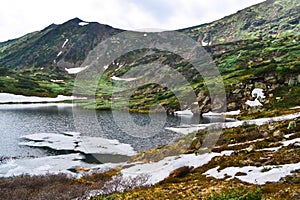 mountain lake among snow glaciers in the summer green mountains overgrown with trees under cloudy sky, Siberia