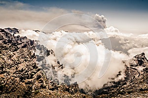 Mountain landscape with clouds and high cliffs. Mountain peaks above the clouds. Cumbre Vieja volcano erupting in the background.