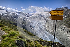 Mountain landscape with green fields and snowy peaks and wild glacier in the Swiss Alps and yellow hiking trail signpost with the