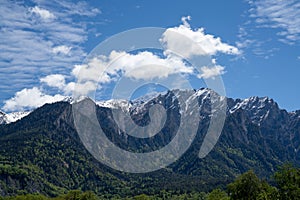 Mountain landscape with jagged snow-covered peaks and green forest under an expressive blue sky