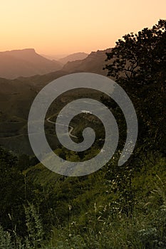 Mountain landscape - panorama view on lush green ridges, folded hills, serpentine road, village away, tree in silhouette in orange