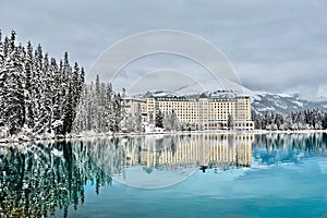 Mountain lodge in winter forest by turquoise  calm alpine lake with snow capped mountains behind it.