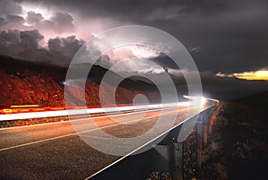 The mountain road with the car passing by the sunset on the right and a thunderstorm with lightning left is shot on a