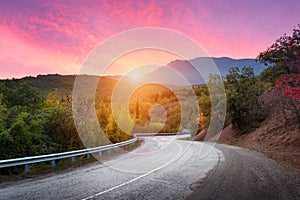 Mountain road passing through the forest with dramatic colorful sky and red clouds at colorful sunset in summer. Mountain